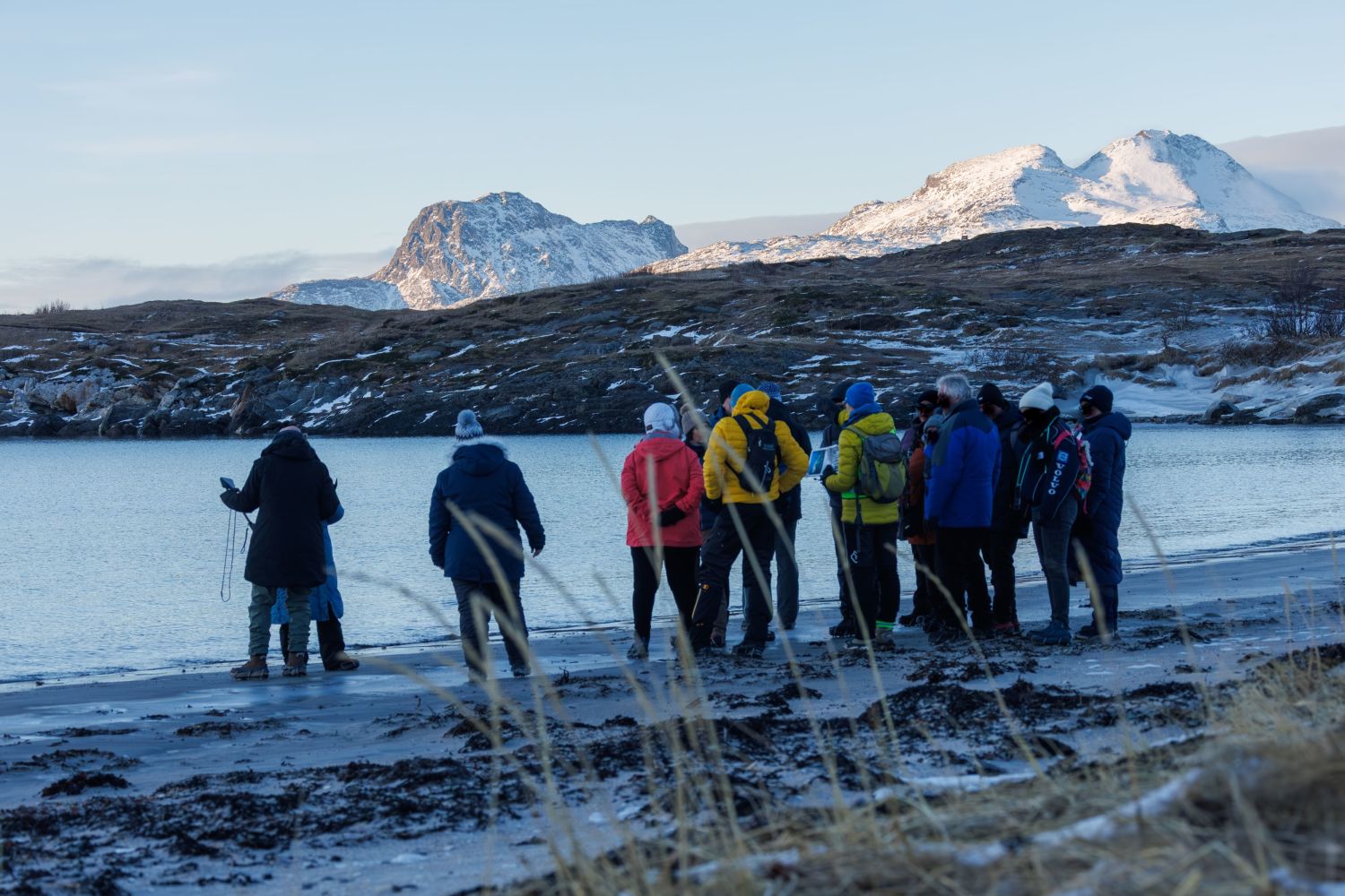 A group of people dressed in winter clothing gathered along a rugged shoreline during an Arctic coastal walk in Bodø, with calm water and snow-covered mountains in the background under a clear sky.