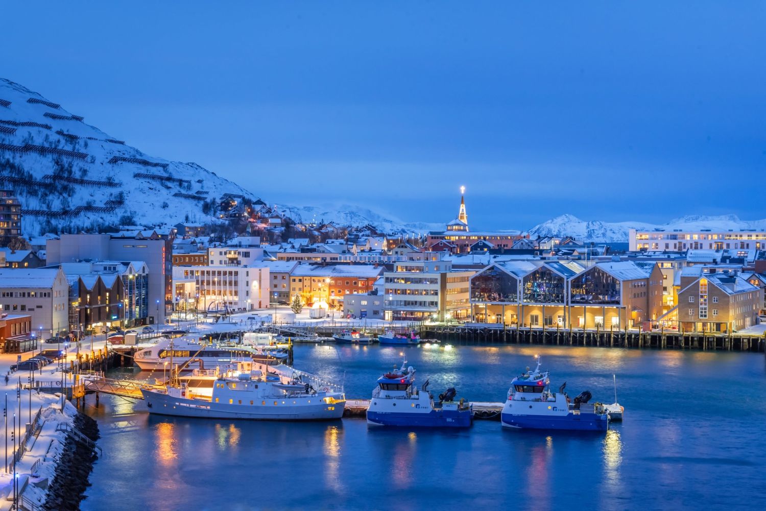 A winter evening view of Hammerfest harbour in Northern Norway, with snow-covered buildings and hills, brightly lit waterfront warehouses and hotels, several docked vessels in the foreground, and a church with an illuminated cross standing above the town.