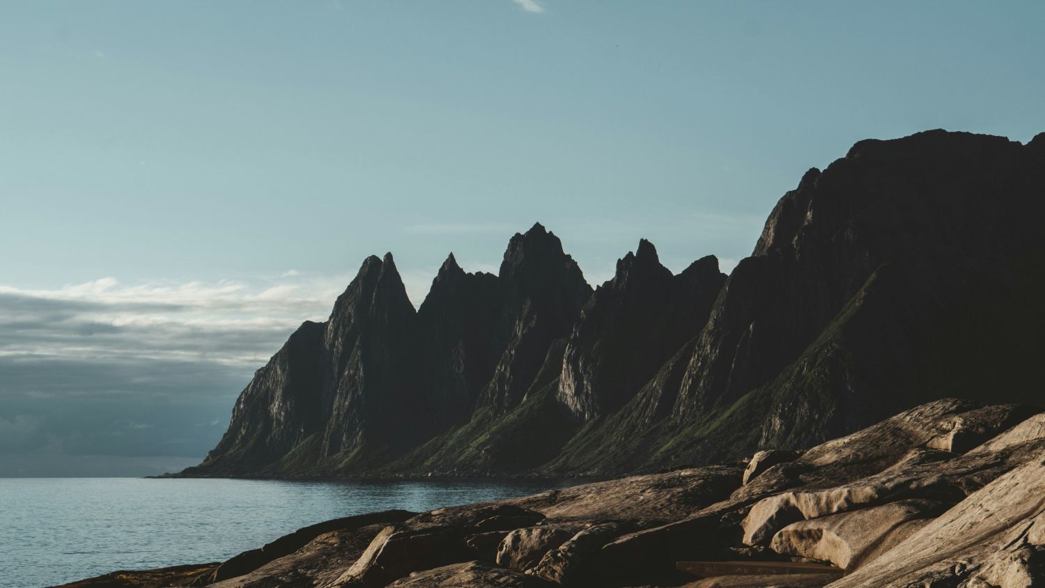 Sharp mountain ridges rise straight from the sea along Senja’s dramatic coastline, their dark faces catching the last, low-angle Arctic light. With bare rock in the foreground and calm water at the edge, the landscape feels ancient, quiet, and endlessly imposing.