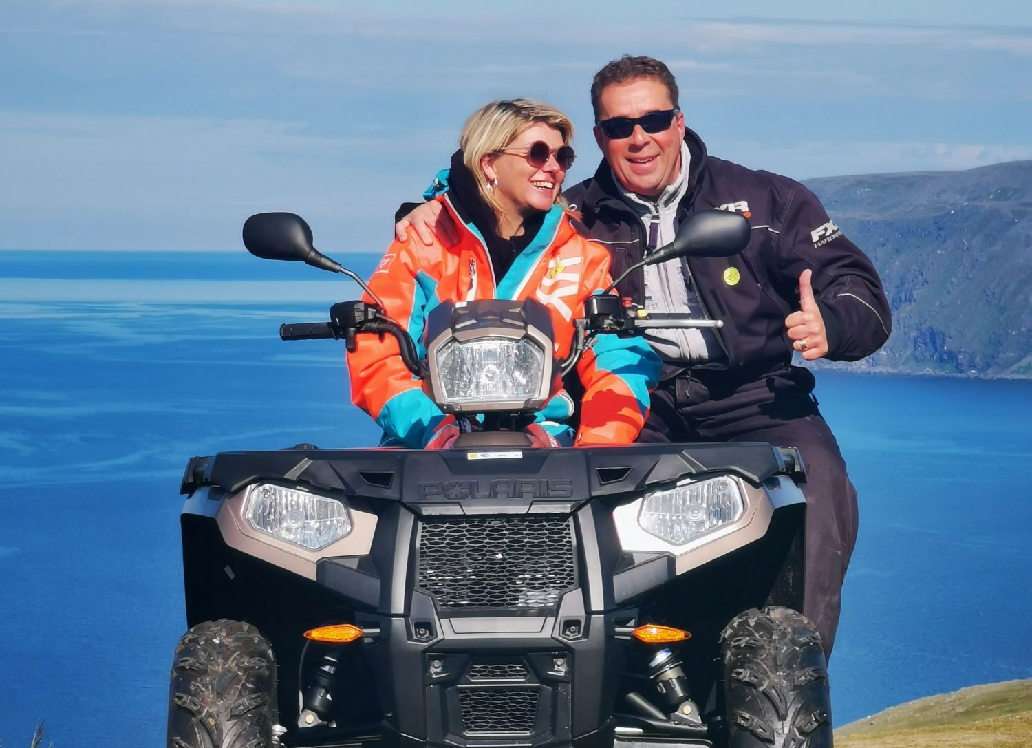 Smiling couple sitting together on an ATV during a North Cape safari, with blue sea and steep cliffs in the background.