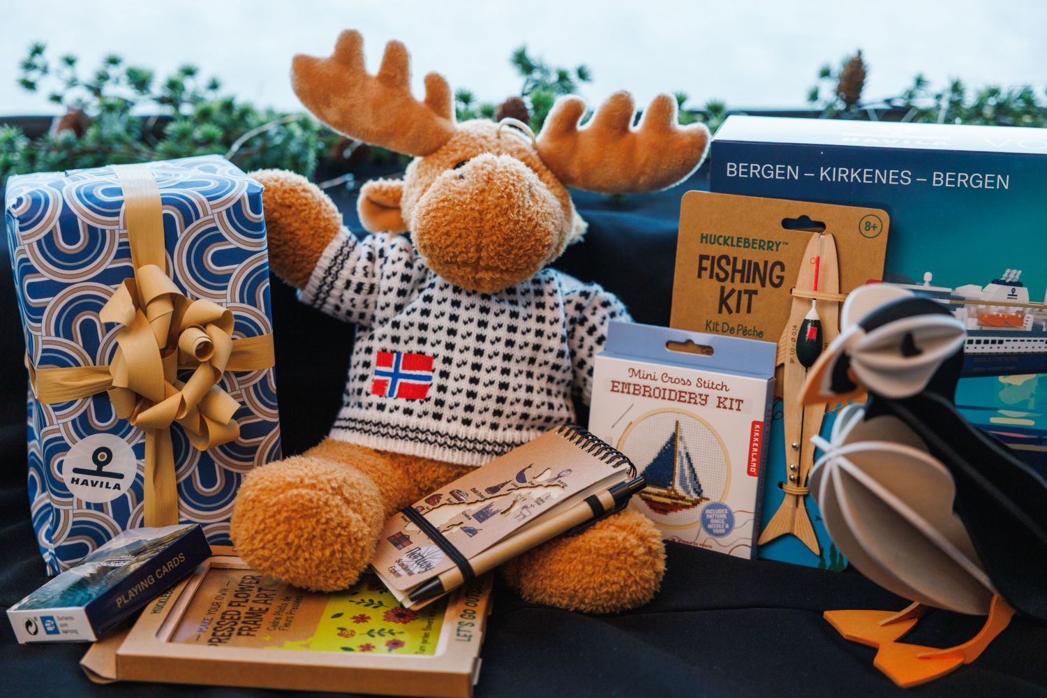 Plush moose in a Norwegian sweater surrounded by Havila gift boxes, notebooks, toys, and souvenirs on a table, with a Bergen–Kirkenes–Bergen route sign in the background.