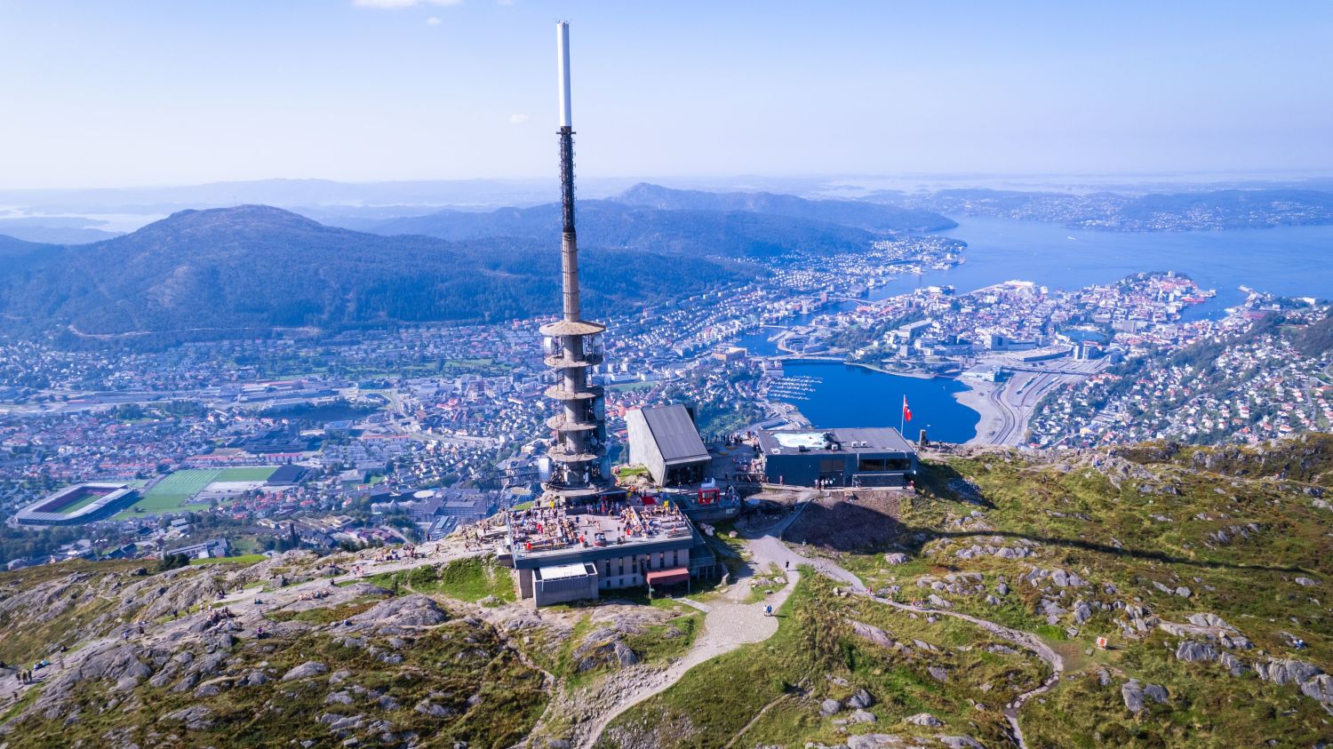 View from Mount Ulriken in Bergen, featuring the cable car top station, the TV tower, and the city of Bergen in the background. Fjords, neighborhoods, and surrounding mountains are visible in the landscape.