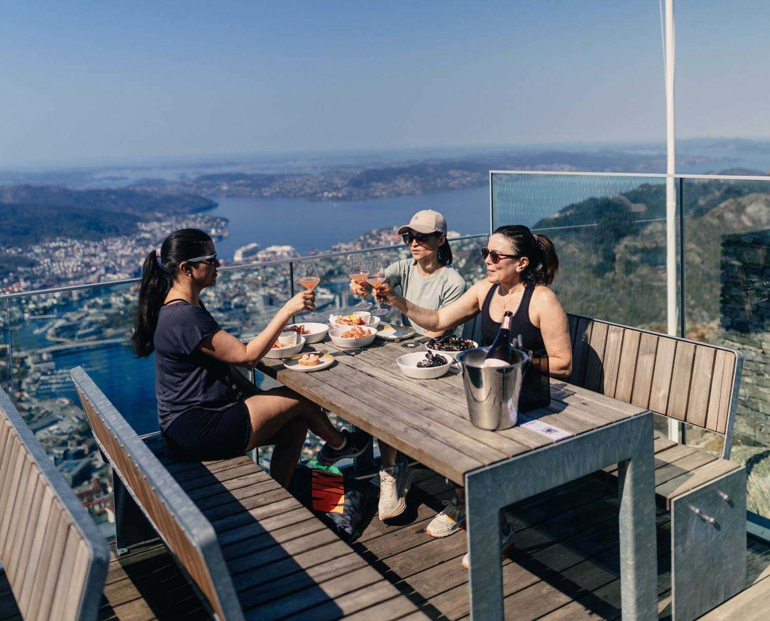 Three women enjoying seafood and drinks at a mountaintop terrace restaurant with panoramic views over Bergen and the Norwegian fjords from Mount Ulriken.
