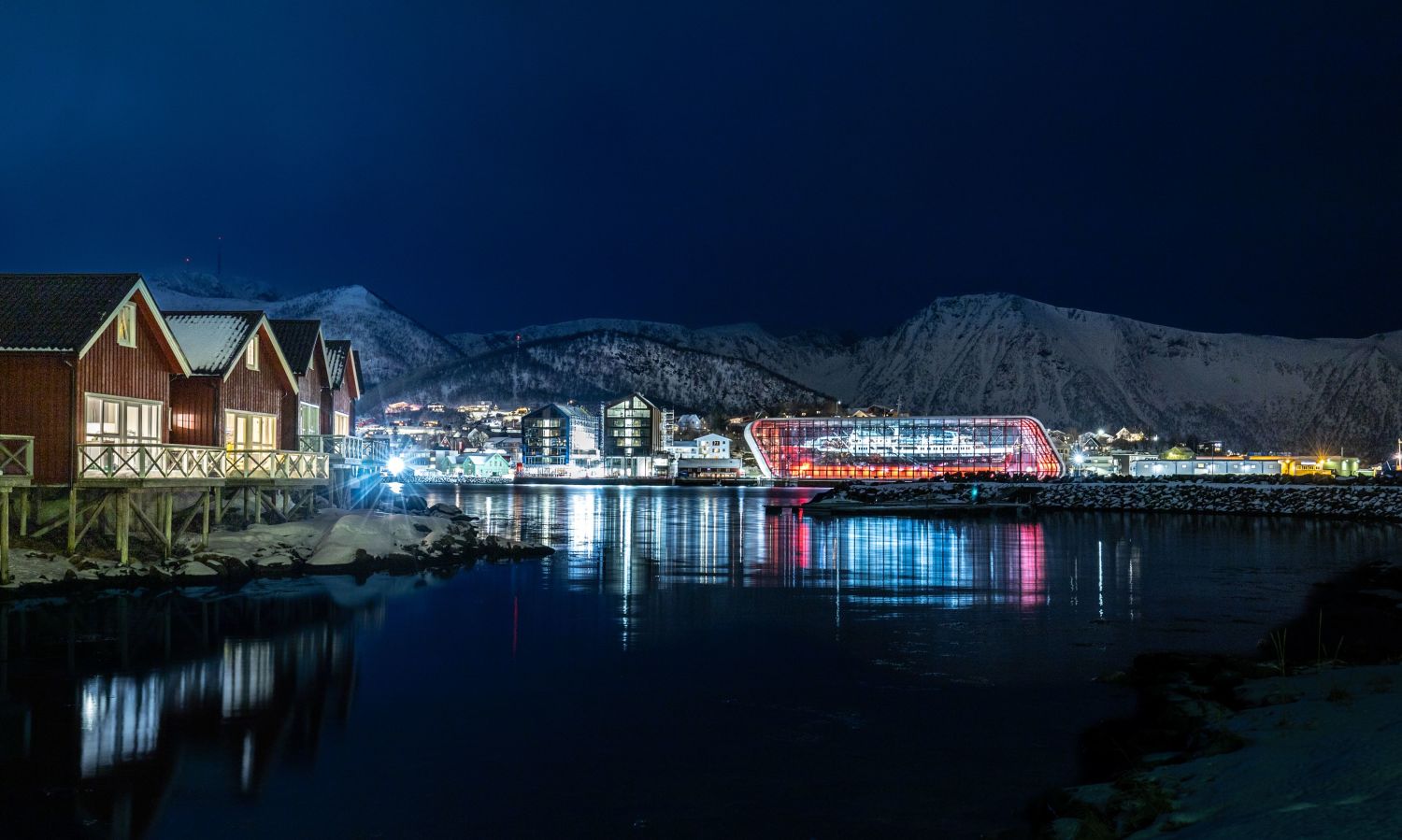 Night view of the Hurtigruten Museum in Stokmarknes, Vesterålen, with the illuminated glass building by the waterfront, snowy mountains in the background, and reflections in the calm harbour.