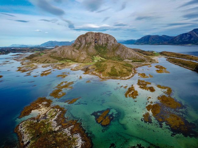 Drone photo of Torghatten near Brønnøysund, surrounded by turquoise shallow waters, skerries, and small islands under a cloudy sky.