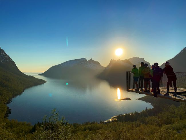 Golden light from the low Arctic sun reflects across a calm fjord on Senja, Norway, as a small group pauses at a mountain viewpoint to take in the vast landscape. Snow-dusted peaks, deep blue water, and fresh spring greenery come together in a quiet moment where nature completely steals the show.