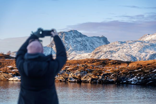 A person in winter clothing taking a photo of snow-covered mountains across calm water during an Arctic coastal walk near Bodø, with soft light illuminating the rugged landscape.