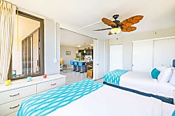 Waikiki Banyan 3401 T2 - bright bedroom with two beds and a tropical ceiling fan overlooking the kitchen area