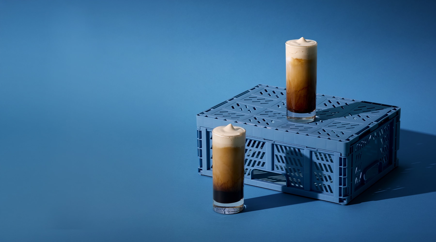 Two glasses of iced coffee topped with cold foam sitting on blue milk crate