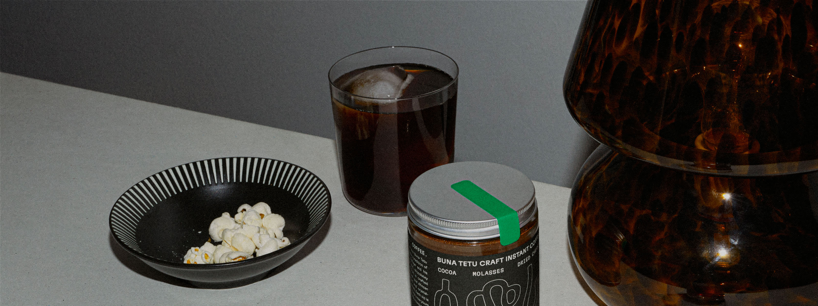 Photograph of Buna Tetu Craft Instant Coffee glass jar next to a prepared instant craft iced coffee in a glass and a small bowl of popcorn