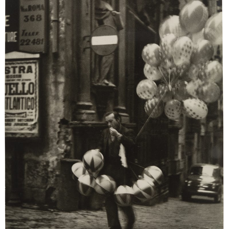 Das Foto zeigt eine schwarz-weiß Fotografie. Das Bild zeigt einen Ballonverkäufer, der auf einer Straße mit Kopfsteinpflaster und italienischen Straßenschildern. In der Hand hat der Mann einen großen Strauß Luftballons und zudem trägt er eine Reihe Ballons um die Hüfte gebunden. Im Hintergrund sieht man einen alten VW-Käfer stehen.