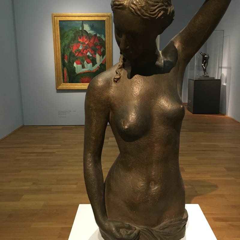 Das Foto zeigt eine Skulptur. Dargestellt ist der nackte Oberkörper einer Frau. Sie legt die Hand in den Nacken und schaut auf den Boden. Im Hintergrund des Bildes sieht man ein buntes Gemälde an der Wand hängen.