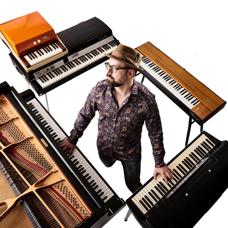 Ein Mann mit Brille und Hut steht in einem Quarree aus Keyboards und Klavieren. 