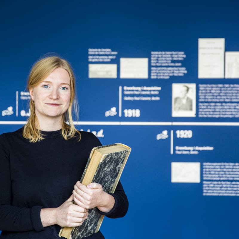 Eine junge Frau mit blonden Haaren steht vor einer blauen Wand mit einer Zeitleiste. In ihrer Hand hält sie ein großes, altes Buch.