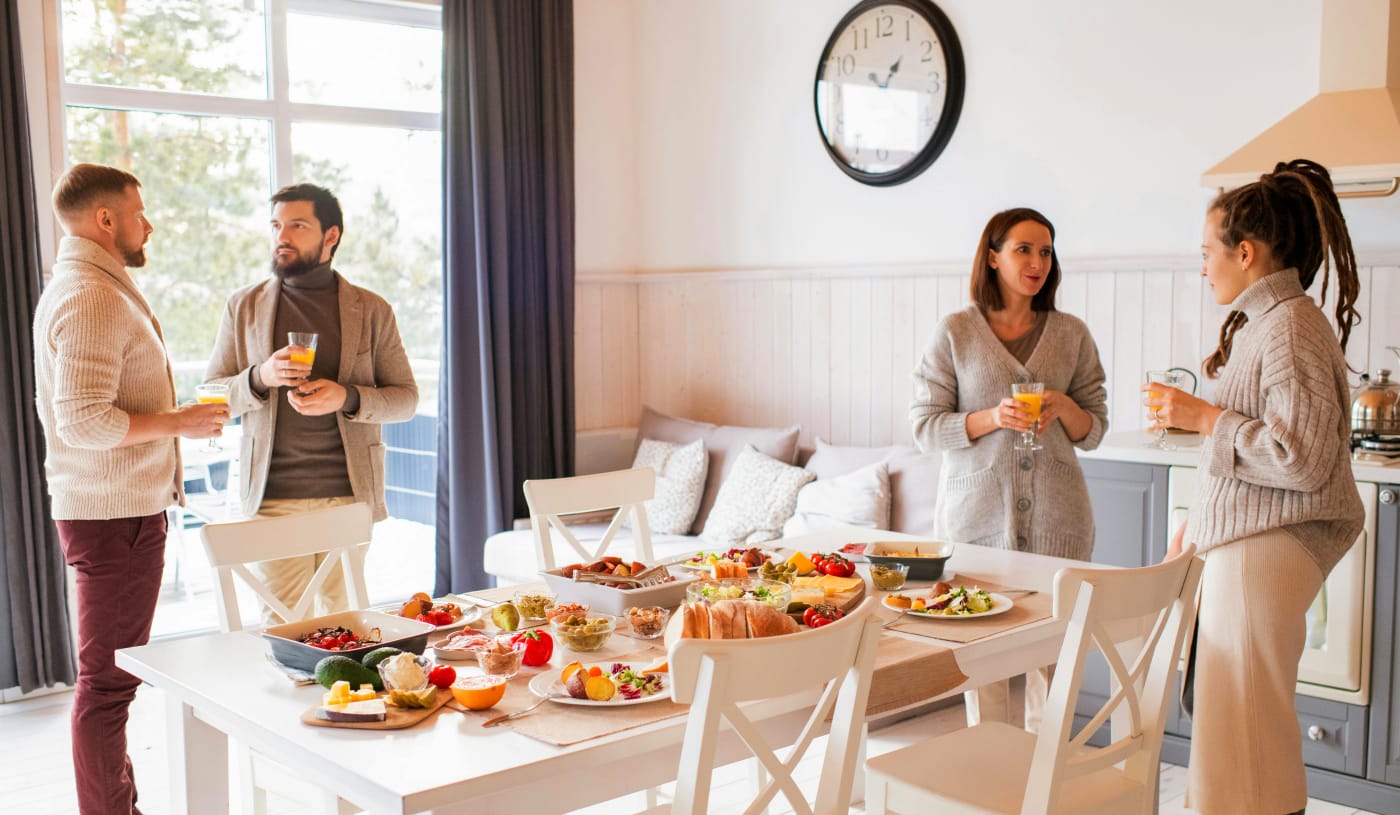 A group of people standing around a table filled with festive snacks and appetizers, socializing in a cozy atmosphere.