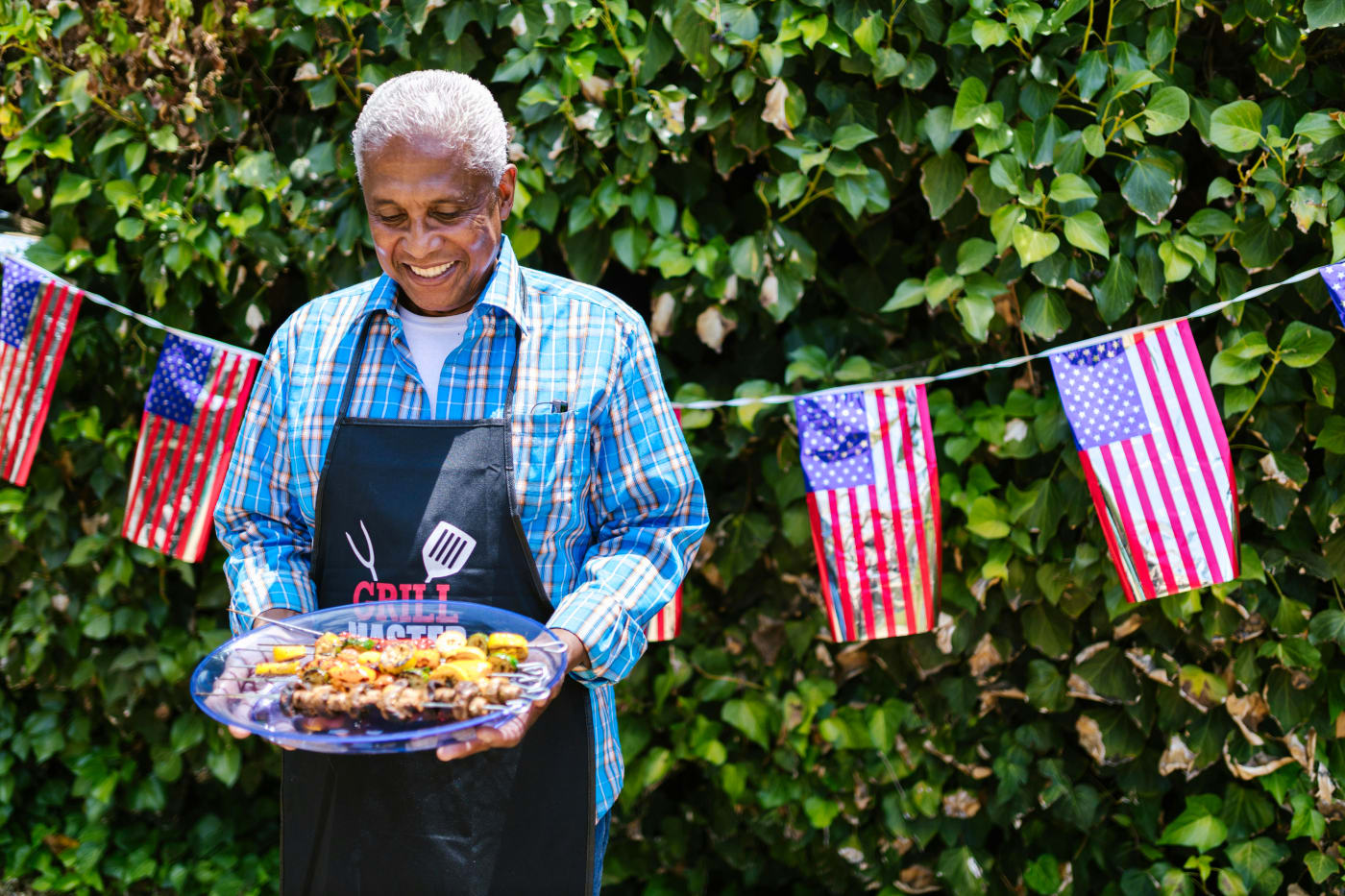 Elderly man in an apron serving American BBQ food at a party with American flags in the background.