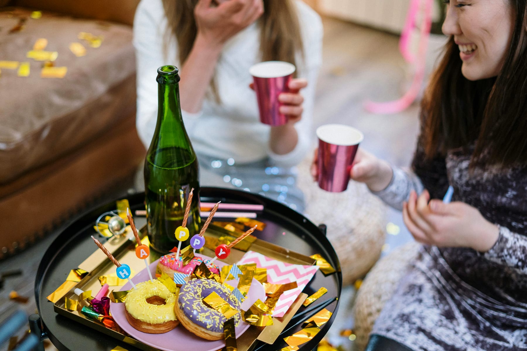 Two women sit at a table filled with party decorations, champagne, and donuts, symbolizing a lively milestone birthday party focused on coziness and decoration.