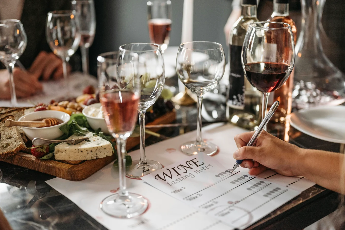 Vinudvalg og madparring: Person skriver noter om vin til forskellige retter med flere glas vin og ostetallerken på bordet.