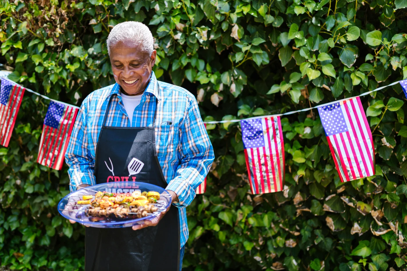 Elderly man in an apron serving American BBQ food at a party with American flags in the background.