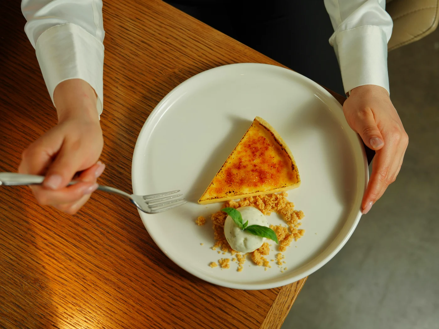 Person enjoying a creme brulee with a perfect caramelized crust, served with garnish and crumbled base.