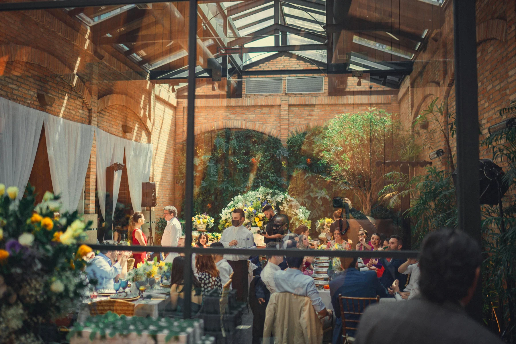 Festive confirmation setup in a beautifully decorated hall with guests sitting at tables, flower arrangements, and a back wall filled with green plants.