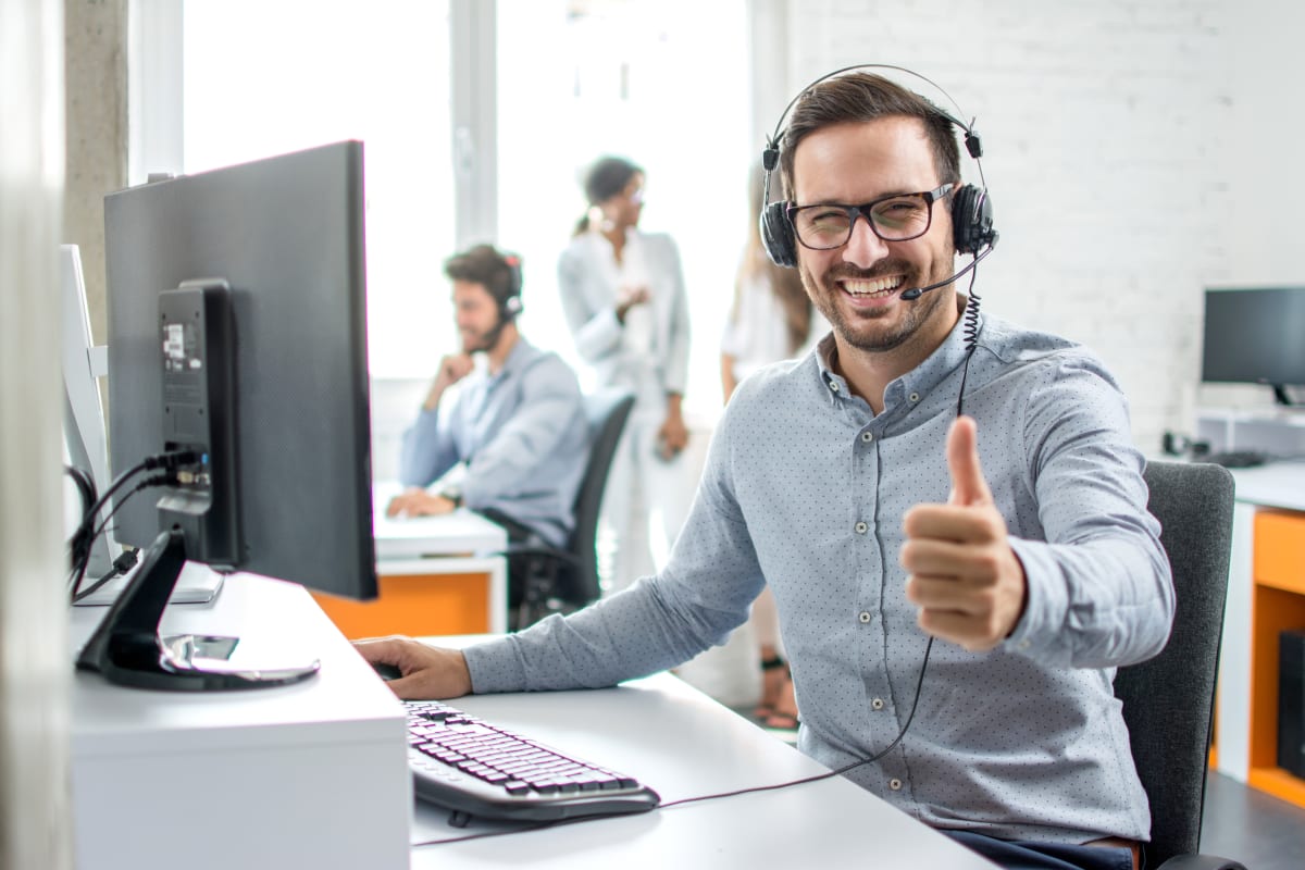 Smiling customer service representative wearing a headset, sitting at a desk with a computer, giving a thumbs-up in a bright modern office with colleagues in the background.