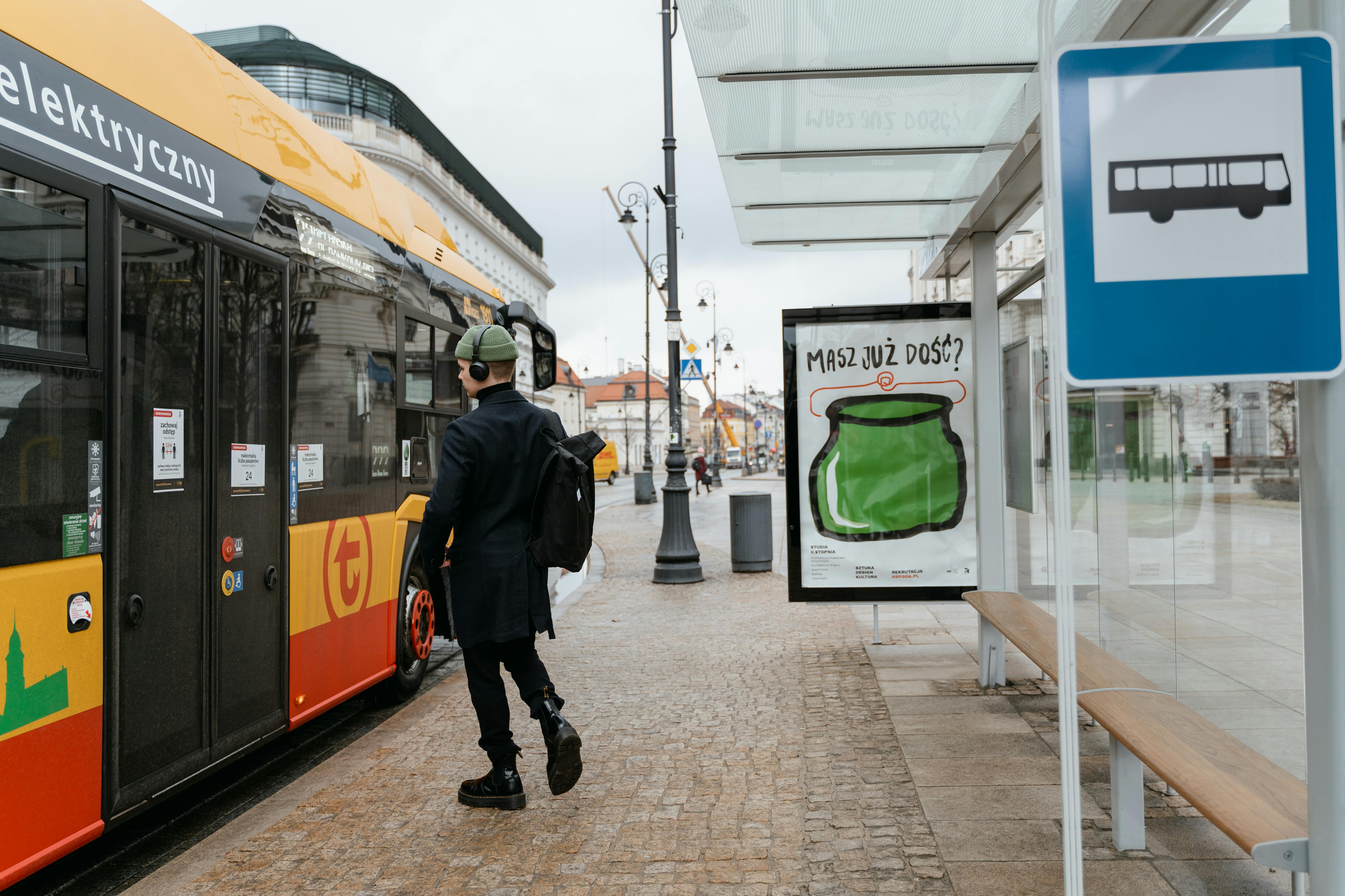 A man with a backpack boards an electric bus at a bus stop on a city sidewalk, showcasing public transportation use.