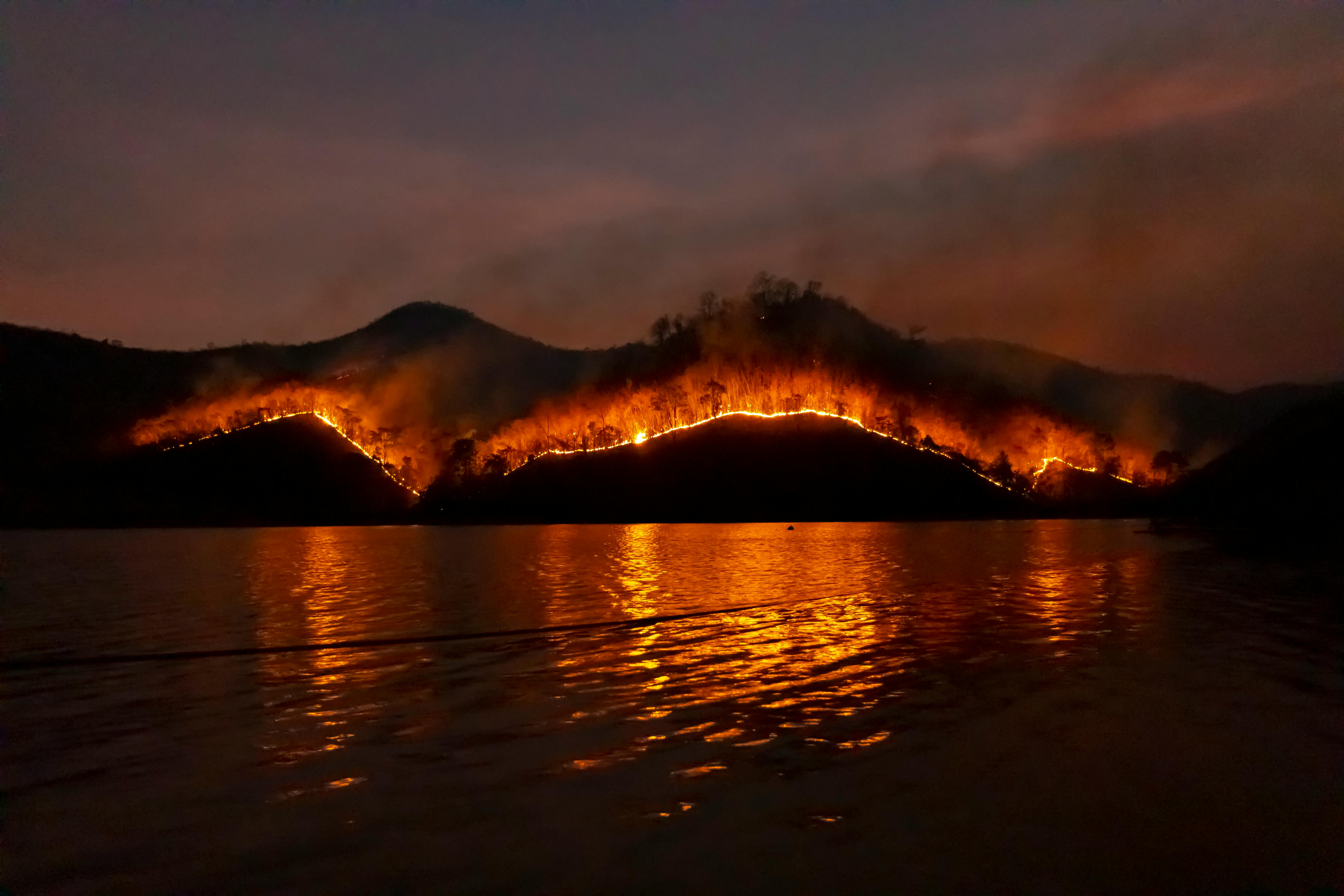Dramatic wildfire in Thailand with blazing mountains reflecting in the dark waters.