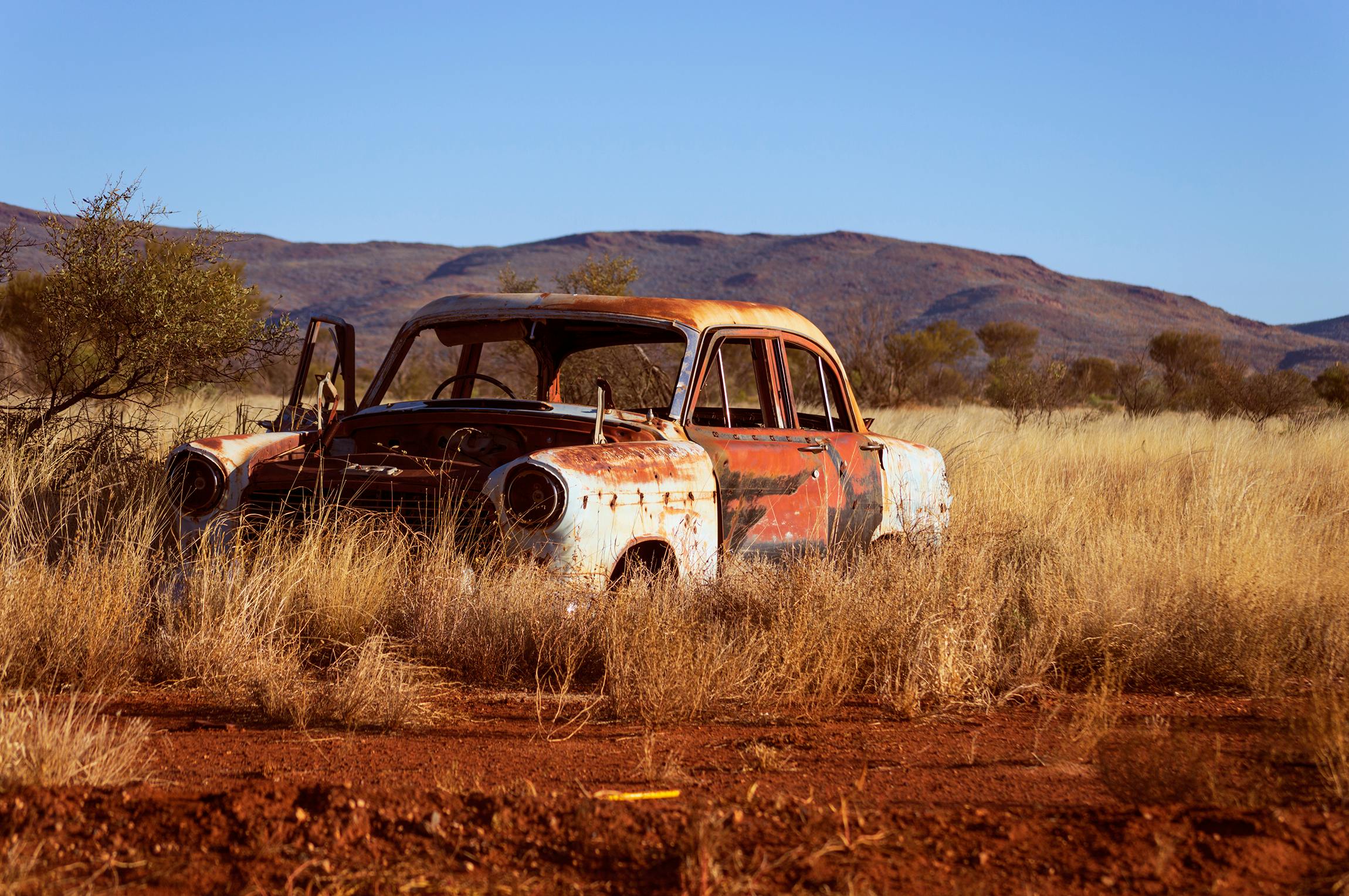 A rusted vintage car sits abandoned in a mountainous desert landscape, embodying decay and time.