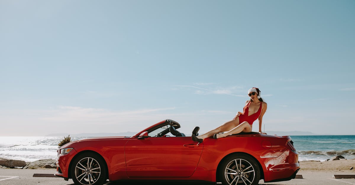 A woman in a red swimsuit lounging on a red convertible by the ocean, enjoying a sunny day.