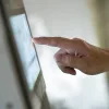 Close-up hand tapping a touchscreen kiosk, finger pressing digital interface.&nbsp;