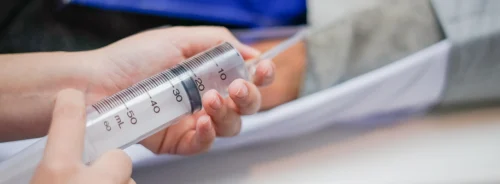 Close-up of hands holding a 60 mL syringe in a clinical setting.