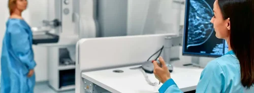 Radiographer at a mammography workstation with a patient at the imaging unit; breast image on screen.&nbsp;