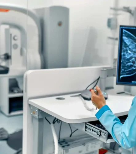 Radiographer at a mammography workstation with a patient at the imaging unit; breast image on screen.&nbsp;