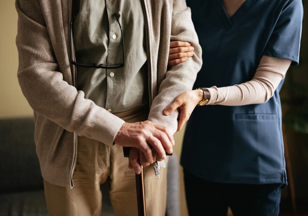 Caregiver supporting elderly man with walking cane during home assistance visit.