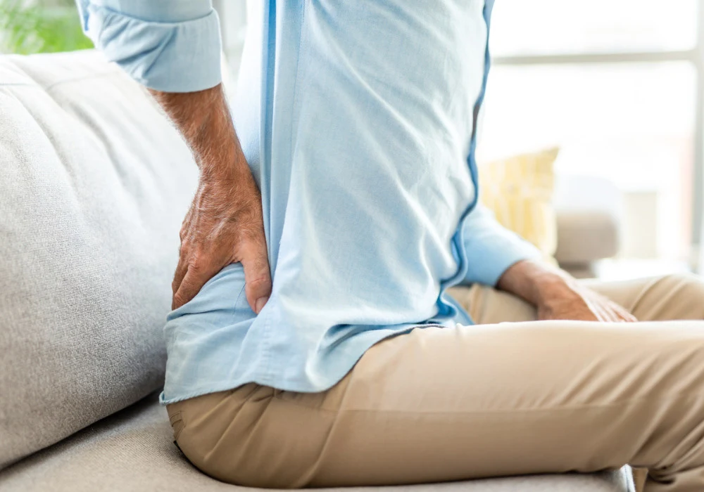 Man suffering from lower back pain while sitting on couch, indicating possible sciatica or spinal issues.&nbsp;
