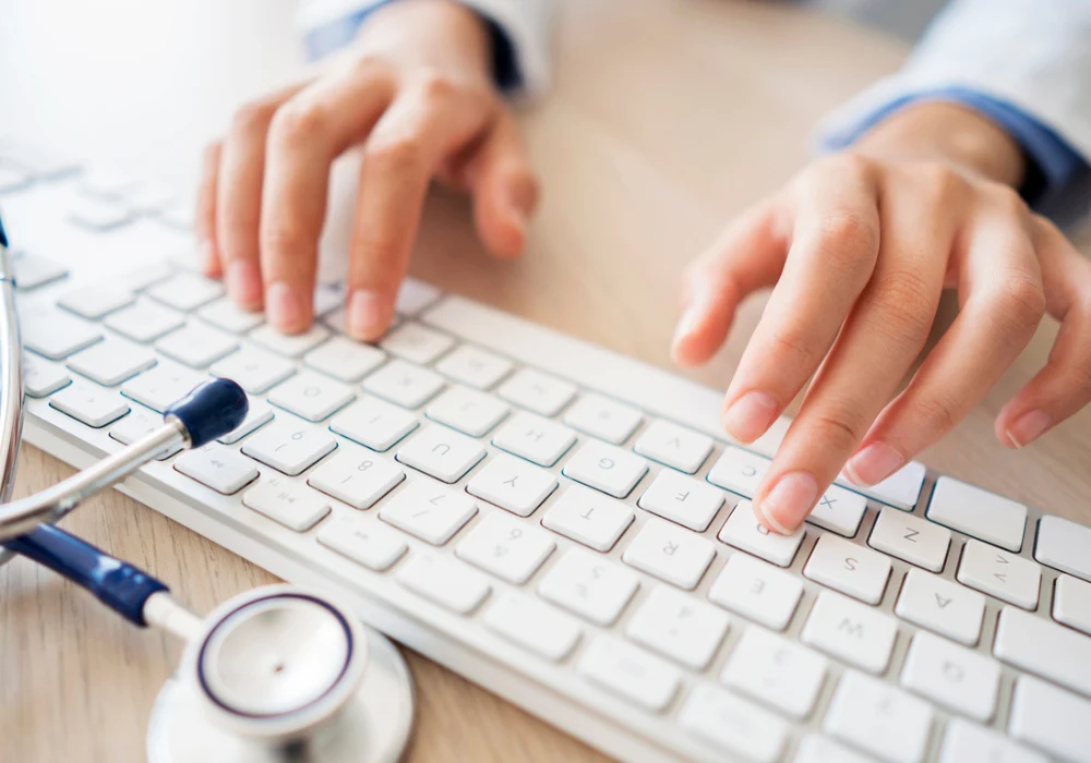 Doctor typing on keyboard with stethoscope nearby, managing electronic health records (EHR)&nbsp;