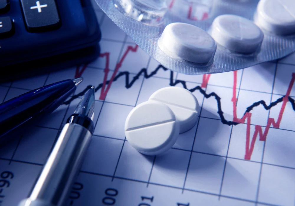 White pills on financial chart with pen and calculator, symbolising rising healthcare costs and pharmaceutical market trends.&nbsp;