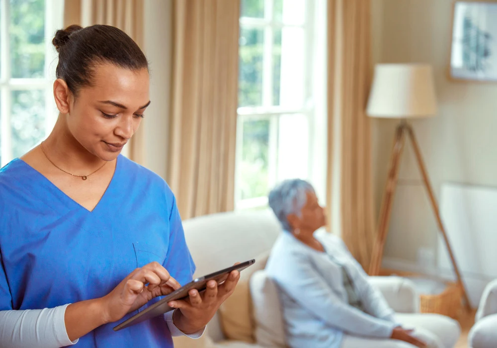 A healthcare worker in scrubs uses a digital tablet while an elderly patient sits in the background.&nbsp;
