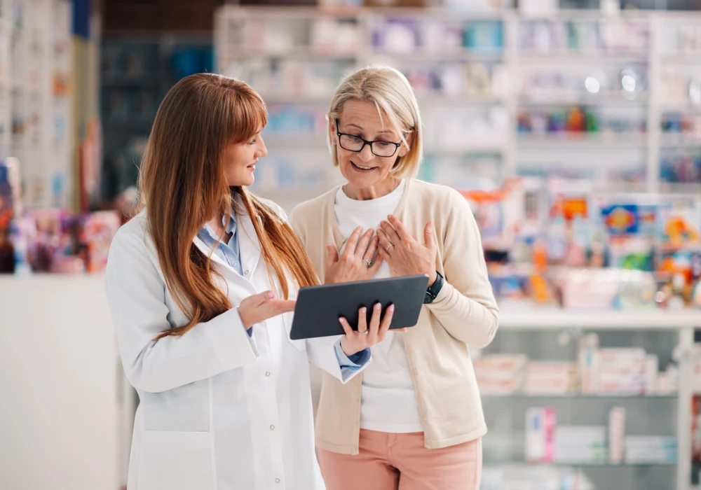 Pharmacist uses a digital tablet to guide a patient in a pharmacy, highlighting pharma-led support, medication access and compliant care.&nbsp;
