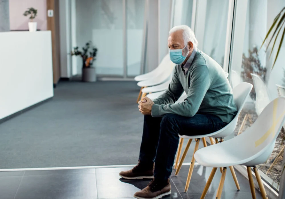 Older man in a face mask sitting in a hospital waiting room during COVID-19, illustrating reduced outpatient visits.&nbsp;