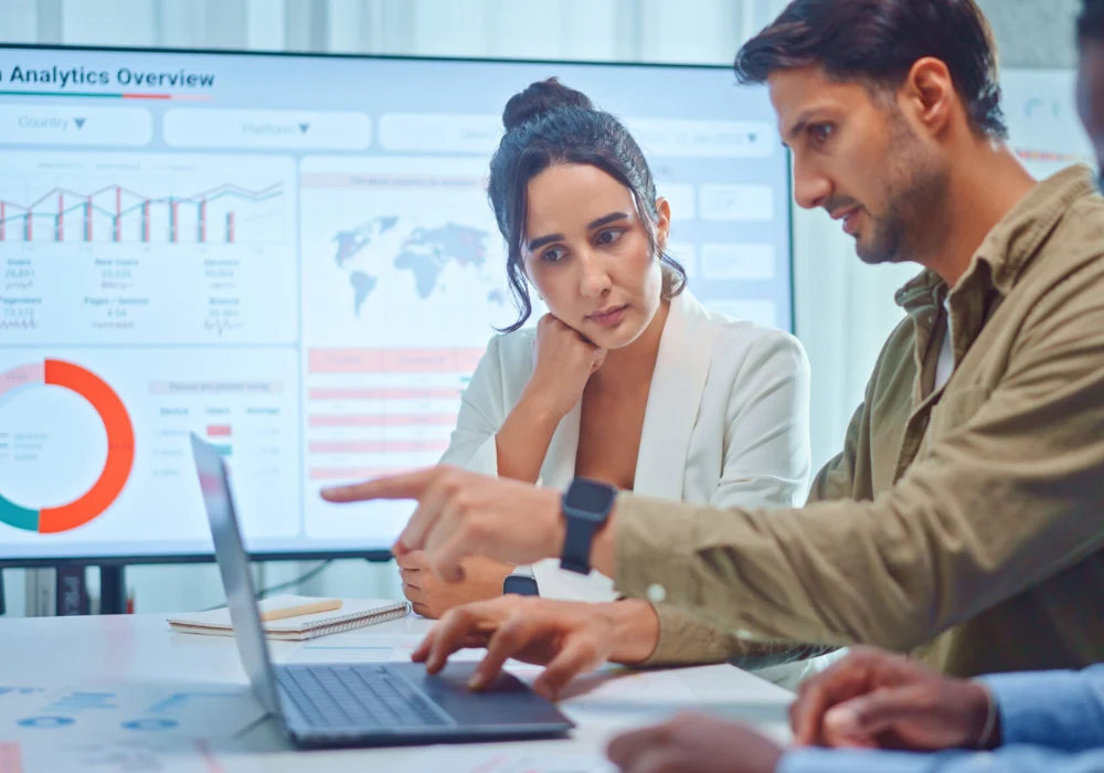 Team of professionals analysing data on a laptop with charts displayed on a screen in the background.&nbsp;