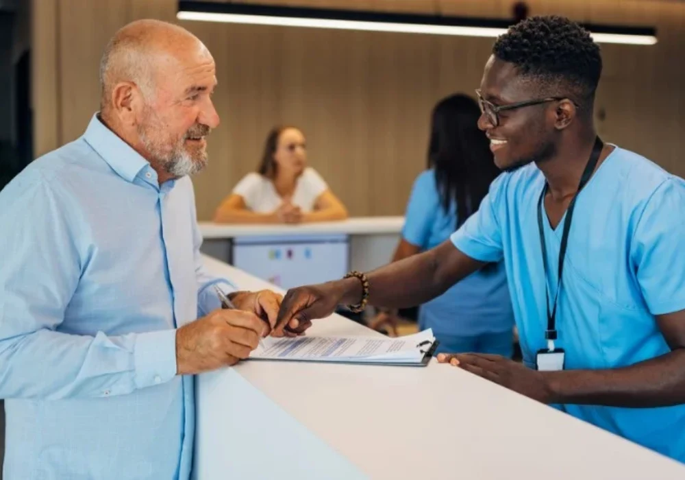 Male nurse assisting an elderly patient at a clinic reception desk, representing patient-centred healthcare and professional communication.&nbsp;