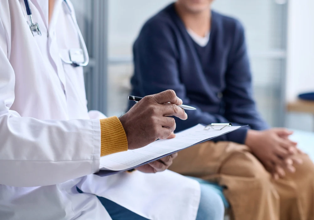 Clinician taking notes during a consultation, patient seated in background.&nbsp;