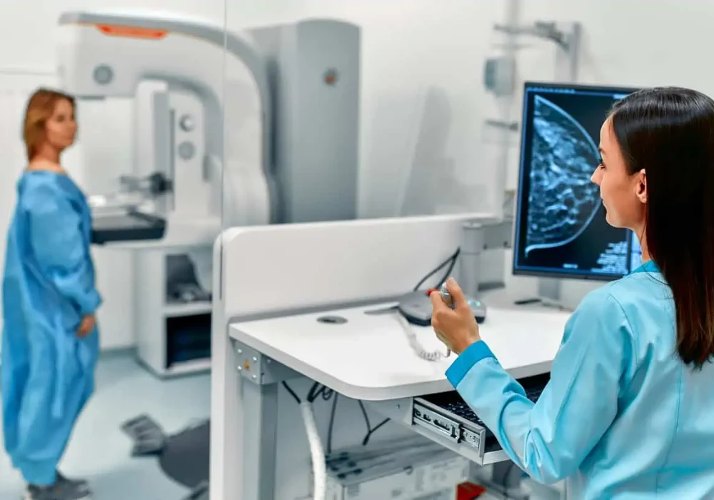 Radiographer at a mammography workstation with a patient at the imaging unit; breast image on screen.&nbsp;