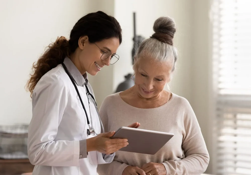 This image shows a healthcare professional (a doctor) with a stethoscope, helping an elderly woman with a tablet,&nbsp;likely discussing&nbsp;her health or medical information.&nbsp;