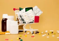 Miniature shopping cart filled with medicine blister packs, surrounded by bottles and scattered pills on a yellow background&nbsp;
