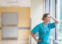 Stressed female nurse looking out hospital window, reflecting emotional burnout and healthcare fatigue.&nbsp;