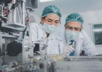  Two scientists wearing lab coats, masks, and hairnets observe pharmaceutical manufacturing equipment.&nbsp;