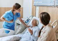 A nurse speaks with a patient lying in a hospital bed while a family member sits&nbsp;beside,&nbsp;depicting patient care, empathy&nbsp;and communication in a healthcare setting.&nbsp;